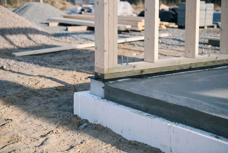 Gjuten betongplatta med vit cellplast i kanten och en syll i trä med nyresta reglar, på grusad mark